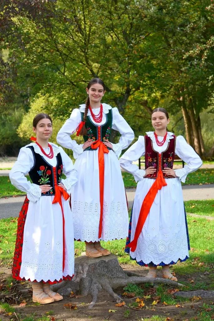 Trzy dziewczyny ubrane w stroje ludowe stoją obok siebie. Są w otoczeniu natury, dziewczyna po środku stoi na ściętym pniu drzewa. Uśmiechają się do kamery.