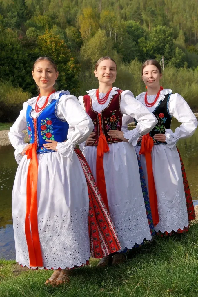 Trzy kobiety w strojach ludowych stoją obok siebie z założonymi na biodrach rękami, uśmiechając się kamery.