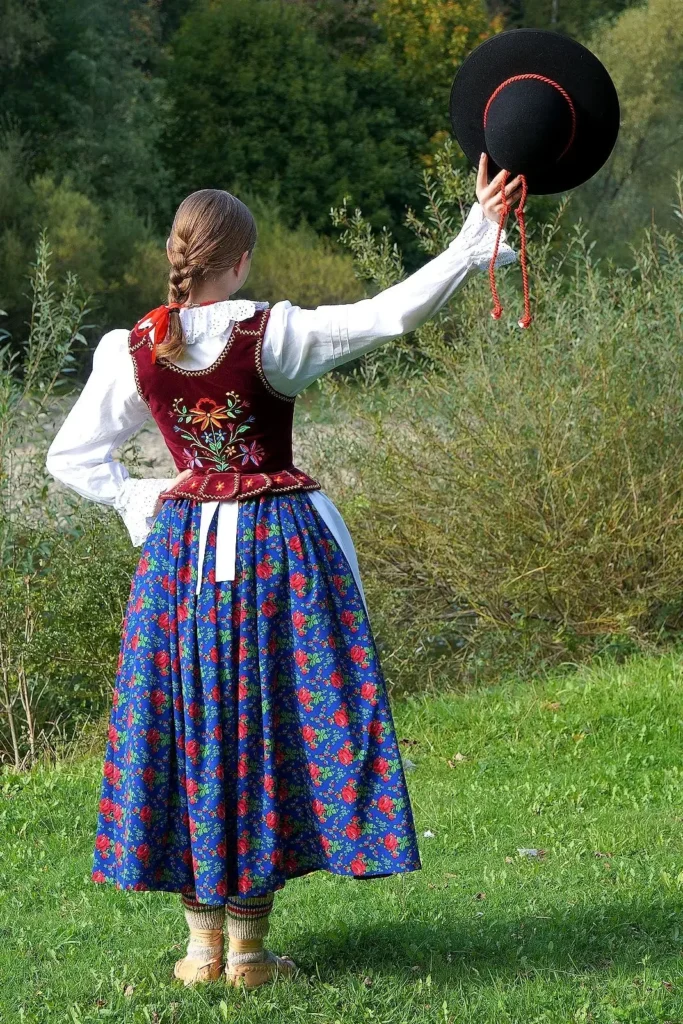 Dziewczyna ubrana w strój ludowy stoi odwrócona tyłem do kamery, unosząc w prawej ręce czarny kapelusz, część stroju ludowego. Znajduję się w otoczeniu natury i pada na nią słońce.