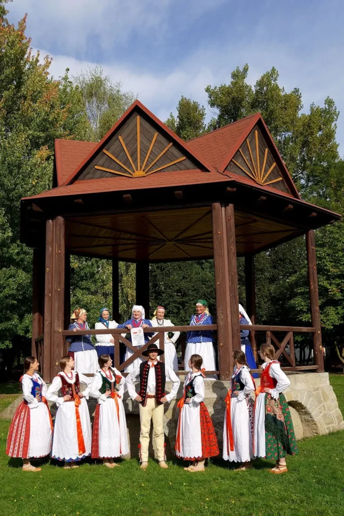 Zdjęcie grupowe z warsztatów tanecznych zespołu ludowego Ziemia Rajczańska. Na trawie stoi sześć młodych kobiet i mężczyzna, w altanie stoją trzy starsze kobiety, wszyscy ubrani są w zdroje ludowe.