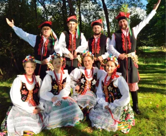 Grupa czterech kobiet klęczących na ziemi, oraz czterech mężczyzn stojących za nimi. Wszyscy mają na sobie krakowskie stroje ludowe.